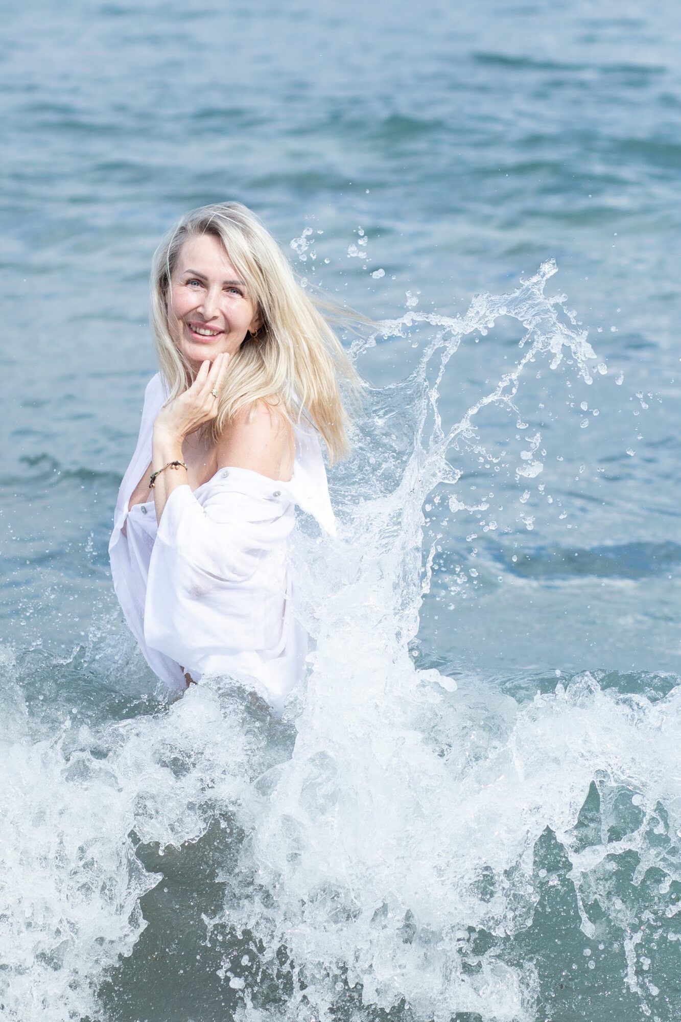Blonde woman in white laughing as a wave splashes around her