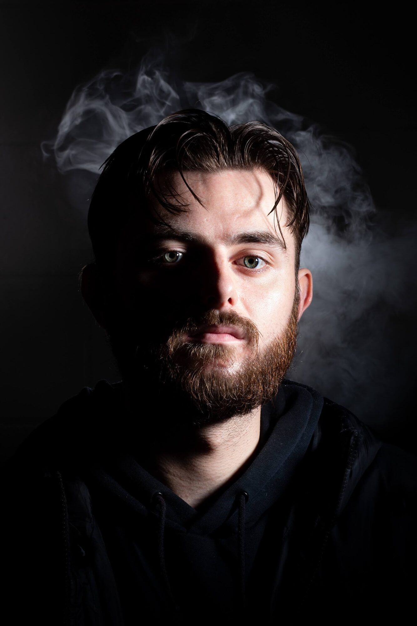 Young man with a dark beard, half his face in shadow, smoke behind