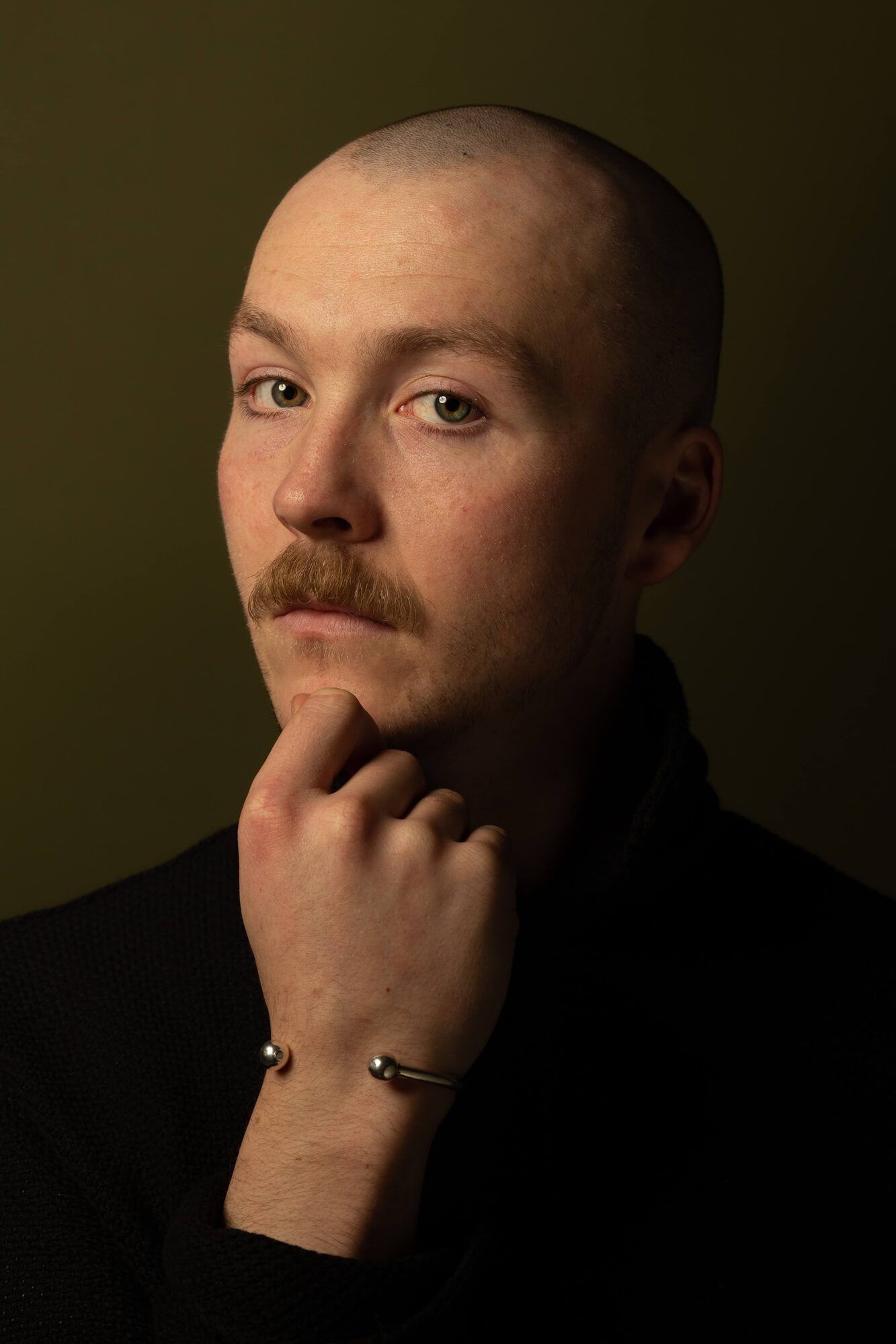 Man with a shaved head and a moustache, hand at his chin