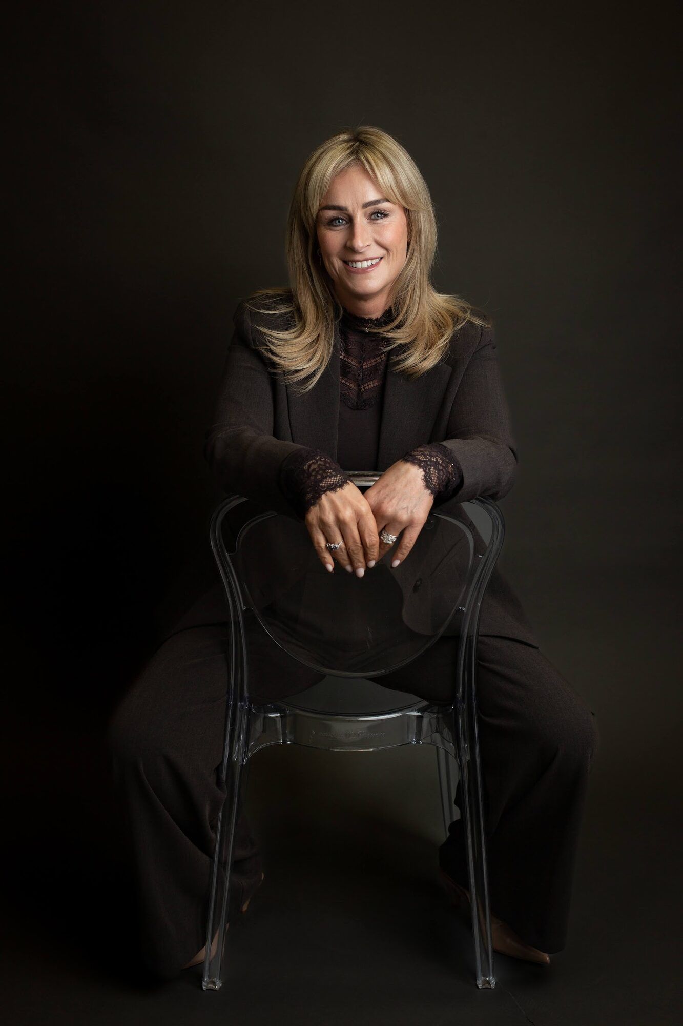 Seated on a chair in black, full length, dark backdrop