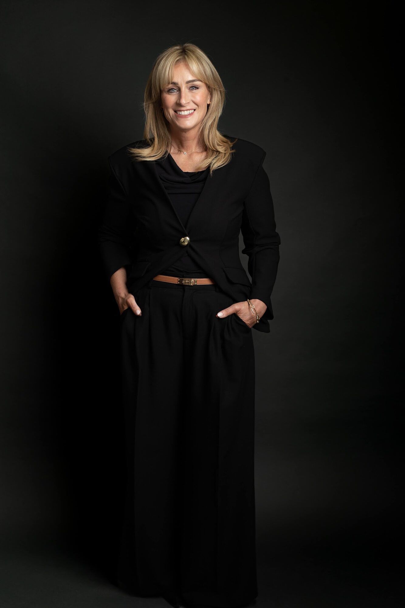 Standing in black, hands in pockets, dark backdrop
