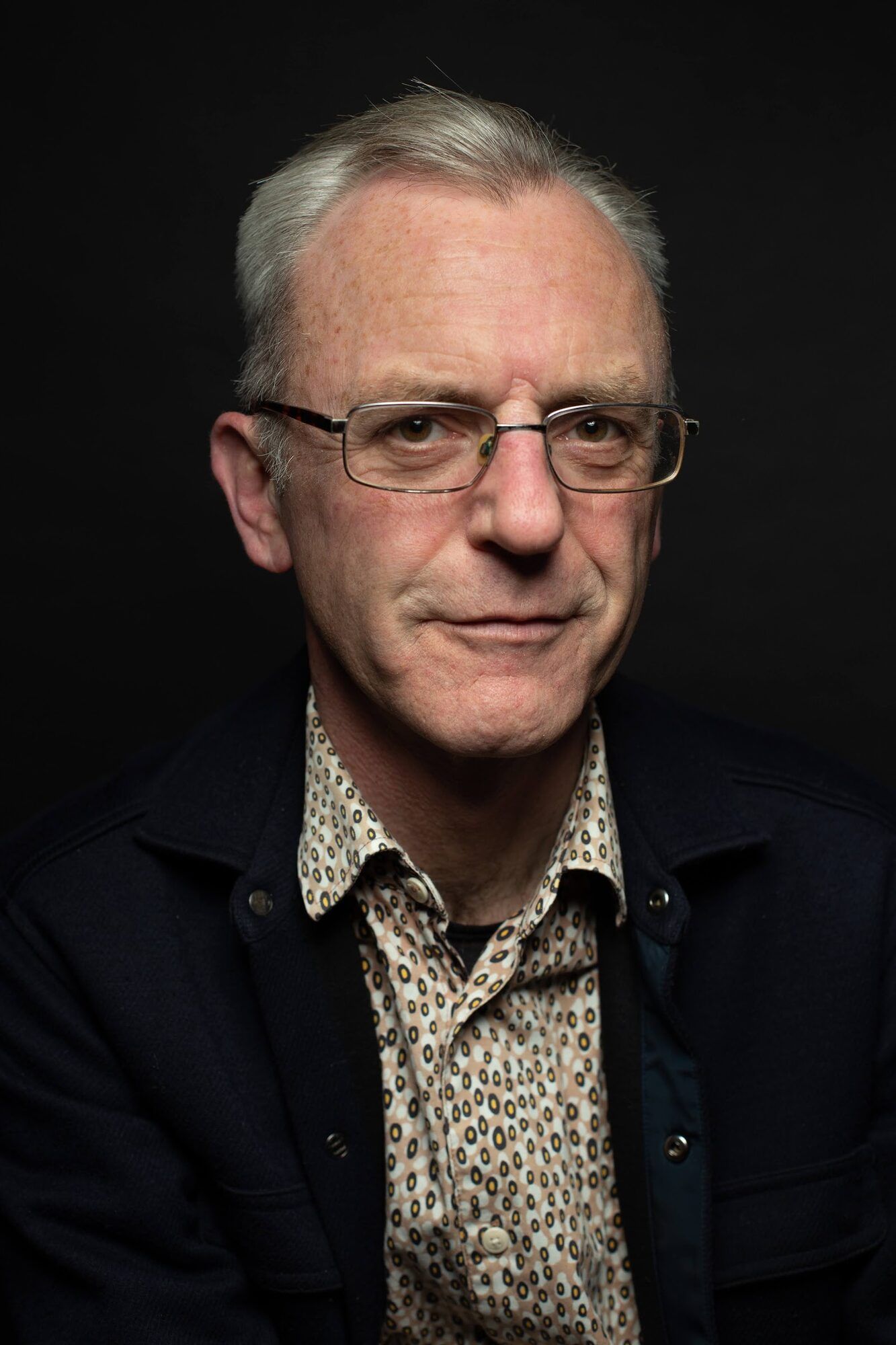 Older man in glasses and a patterned shirt, dark backdrop