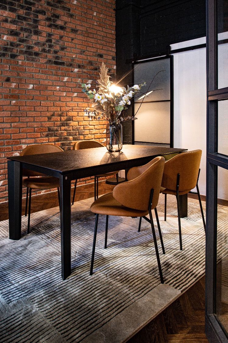 Dining area with a black table, four tan upholstered chairs and a vase of flowers against a brick wall, in warm light