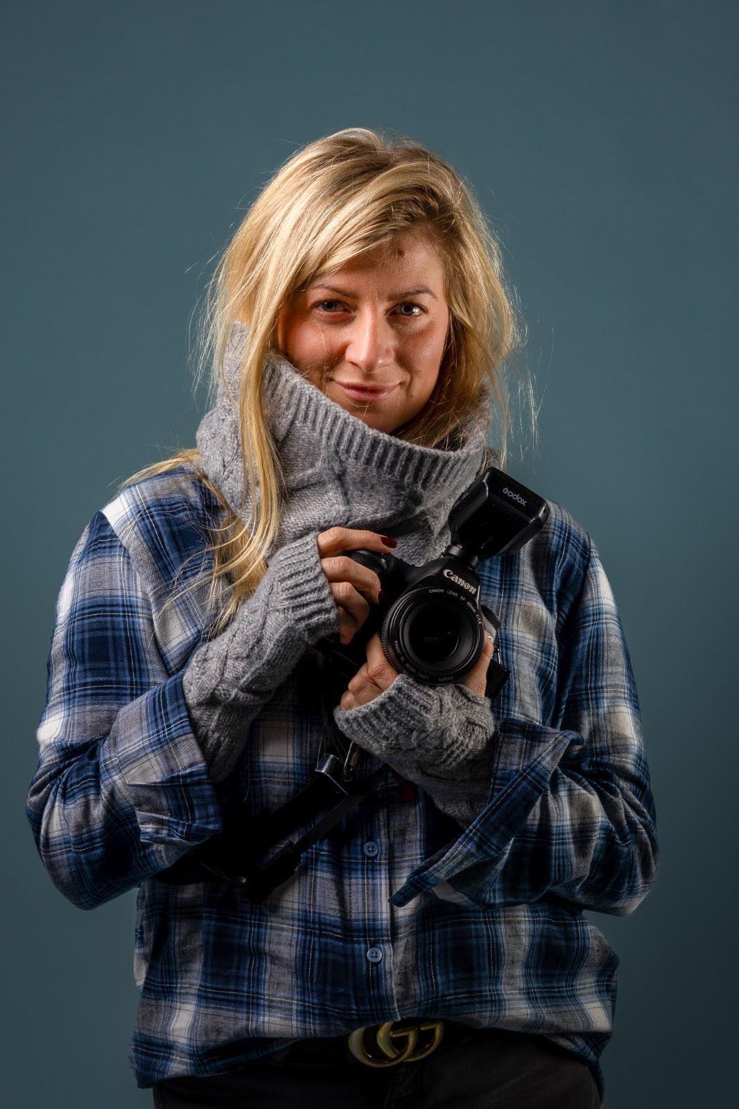 Jadwiga holding her camera, in a checked shirt and grey knitted scarf, against a blue backdrop.